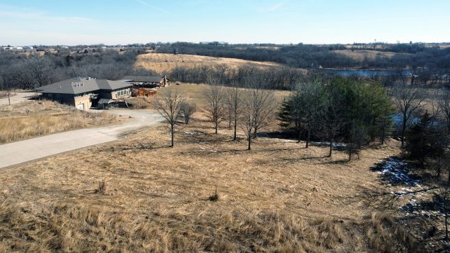 Aerial view of rolling prairie with leafless trees, a nature center building, and a winding paved path under a clear blue sky.